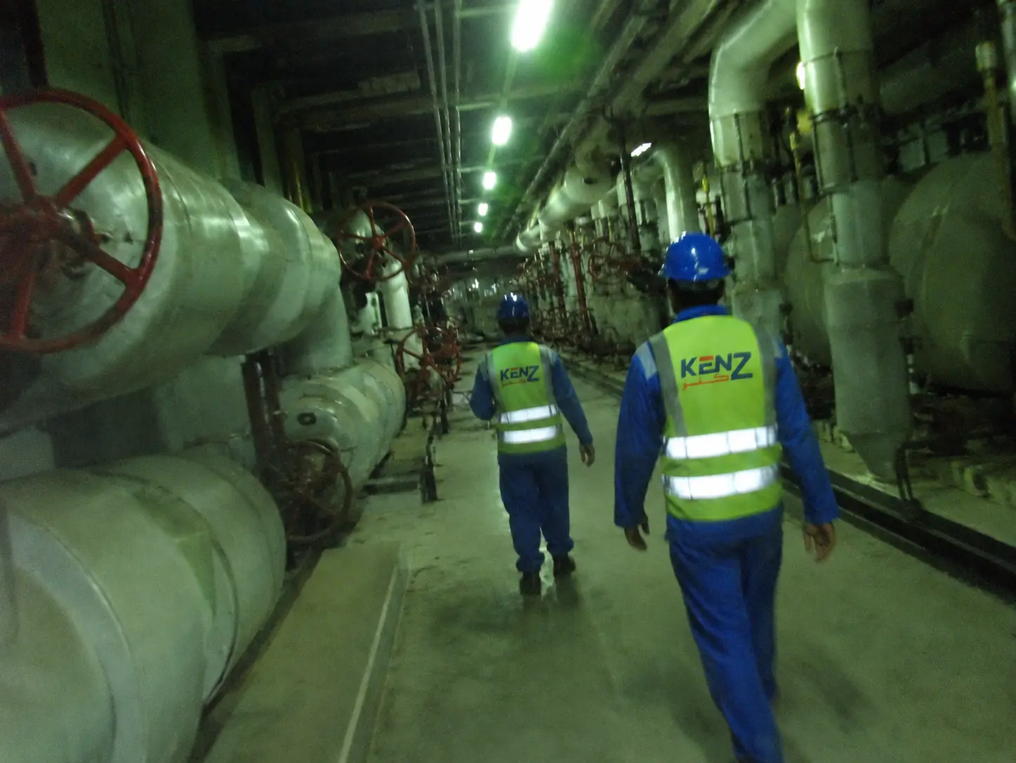 Industrial workers walking through massive underground mechanical plant room with chilled water pipes
