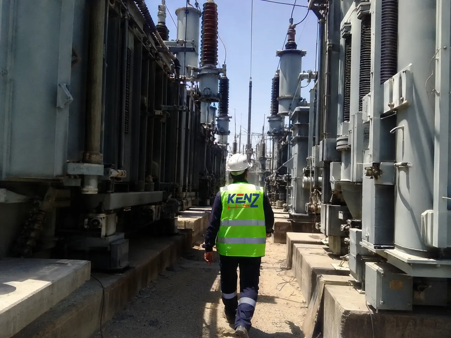 Kenz electrical engineer walking through massive outdoor high-voltage substation with towering transformers in Saudi Arabia